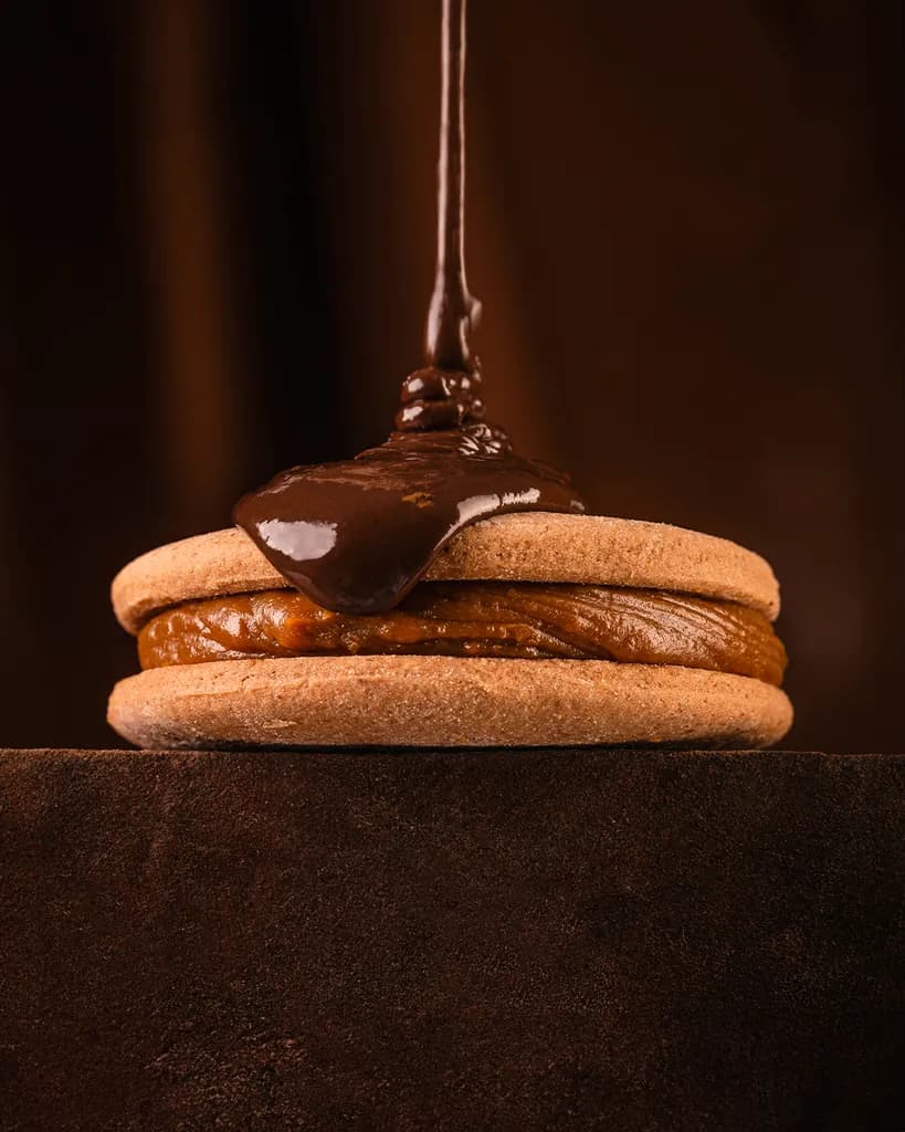 Chocolate derretido cayendo en un hilo sobre un alfajor Malfatti relleno de dulce de leche, sobre un fondo oscuro.