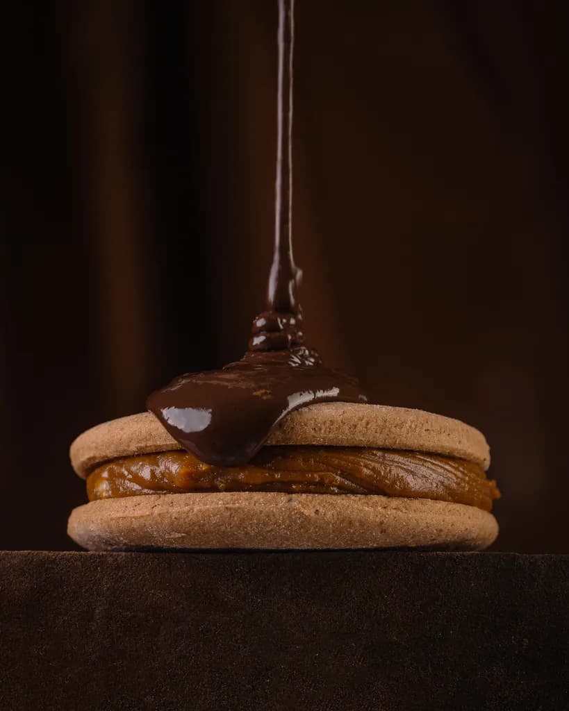 Primer plano de un alfajor Malfatti relleno de dulce de leche: un hilo fino de chocolate le cae en el centro de la tapa y se empieza a extender.