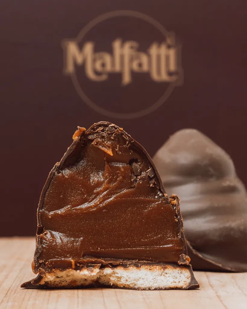 Un Conotte Malfatti cortado al medio, con la cubierta de chocolate y el relleno a la vista, delante del logo de la marca.