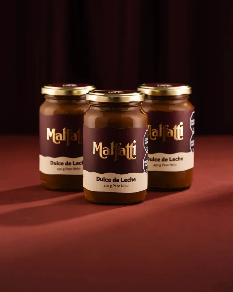 Tres frascos de dulce de leche Malfatti con etiqueta bordó y tapa dorada sobre un paño rojo.