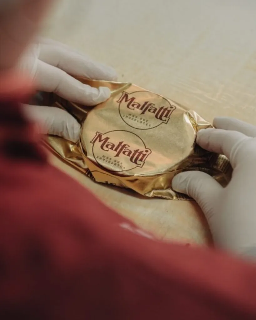 Manos con guantes envuelven un alfajor Malfatti en papel dorado con el logo de la marca.